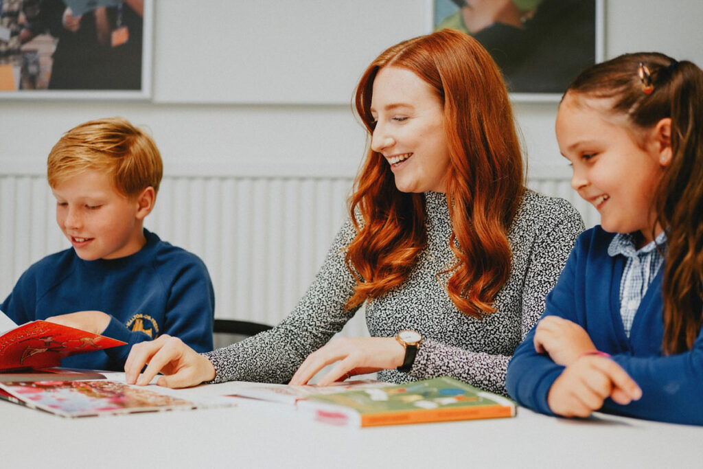 Trainee teacher sat with two students, one either side of her, looking at books with them.