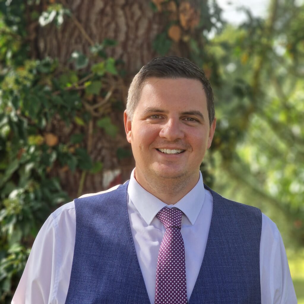 Mike Simmons, Director of JTSCITT headshot. He is stood outside in front of greenery and is wearing a navy waistcoat with a purple tie.