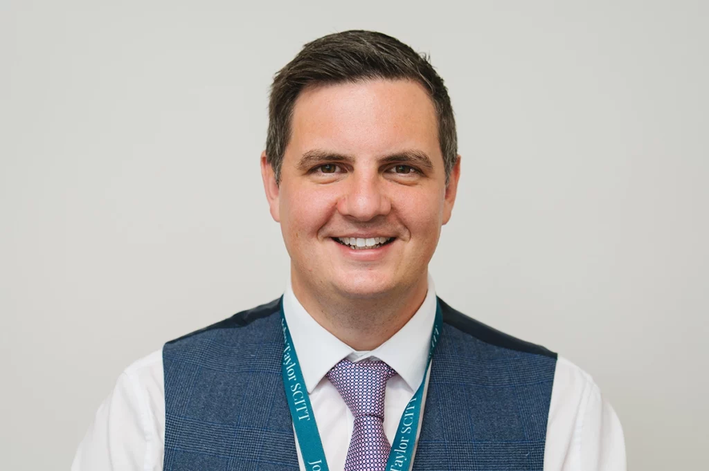 Mike Simmons, Director of JTSCITT headshot. He is stood outside in front of greenery and is wearing a navy waistcoat with a purple tie.